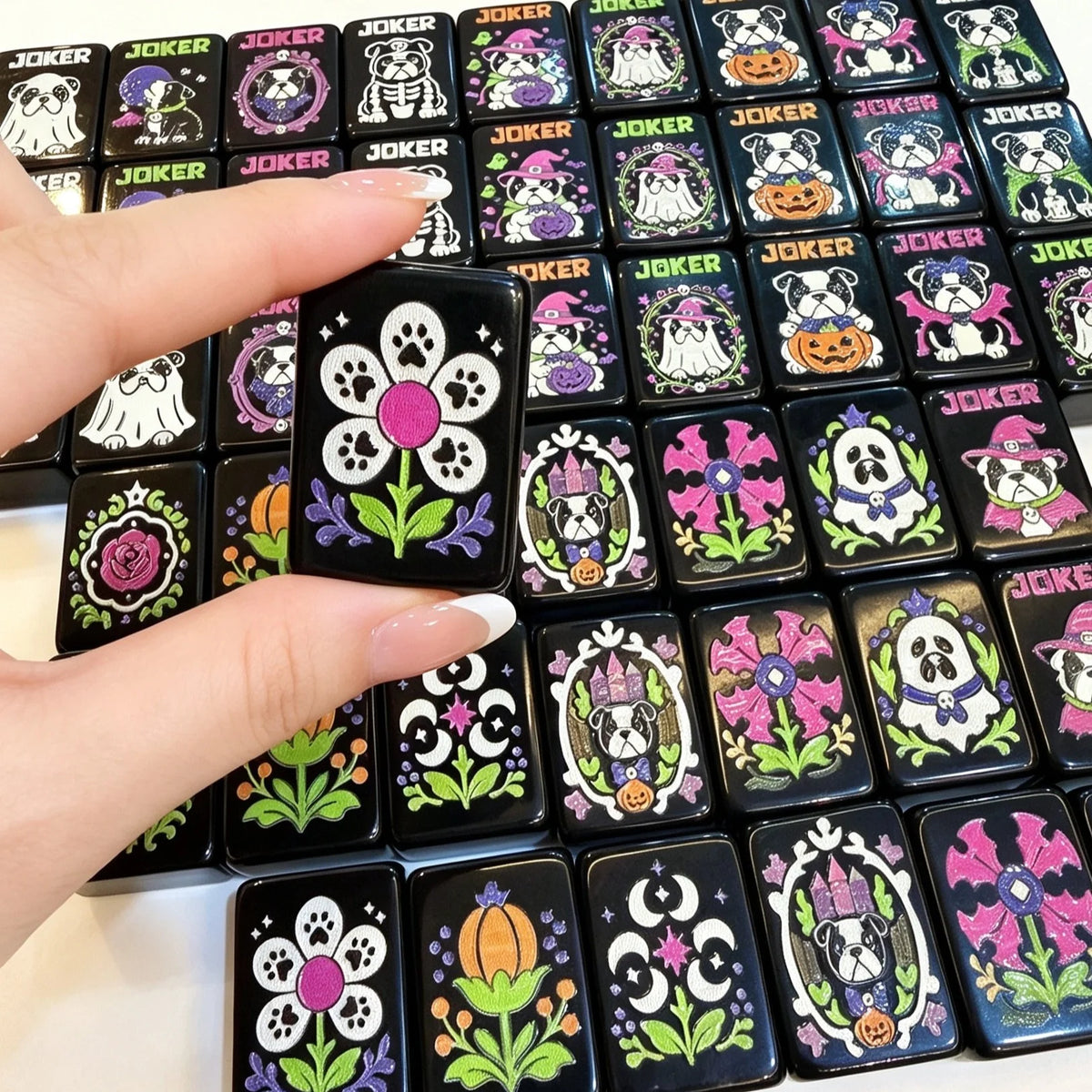 Hand holding a black Mahjong flower tile above Halloween bulldog jokers and illustrated tiles